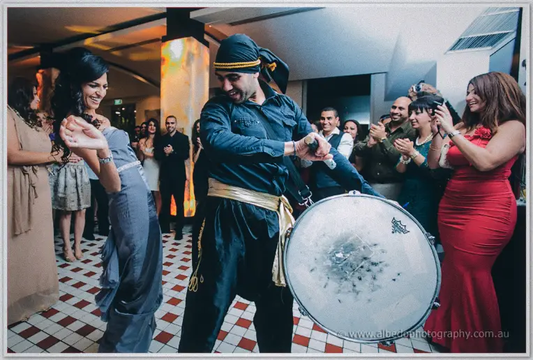 Lydia dances before the drummer in his embroidered sash as clapping guests circle the floor