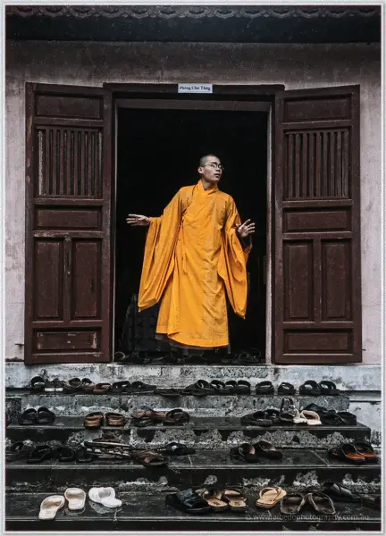 A saffron-robed monk pauses in a crimson temple doorway, shoes lined neatly on the steps