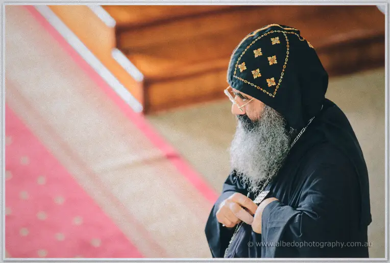 A white-bearded Coptic priest in a black embroidered hood gazes down during the engagement service