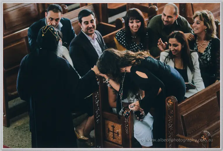 A woman bows to kiss the priest's hand as smiling family members watch from the wooden pews