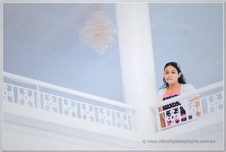 A young girl in polka dots pauses on the white upper balcony beneath a crystal chandelier