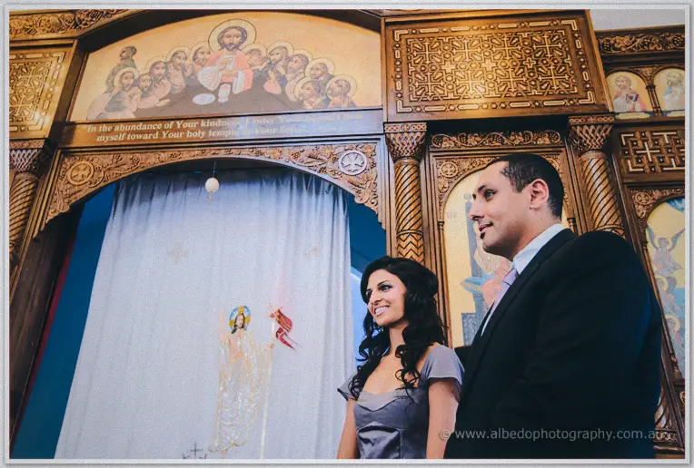 Lydia smiles up at the iconostasis mural while Mina watches on inside the church