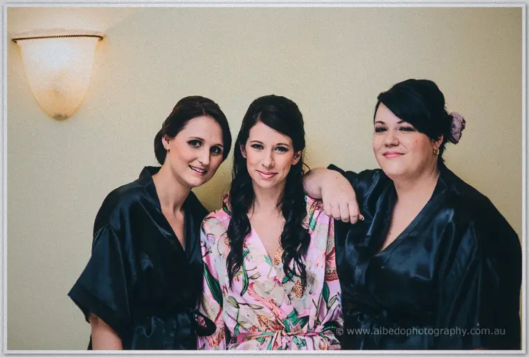 Sarah and her two bridesmaids in black satin robes laugh together against a cream wall at Brookleigh.