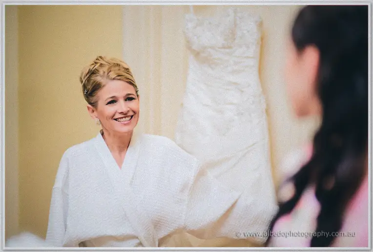 A smiling older woman in a white robe stands beside Sarah's hanging lace wedding gown.
