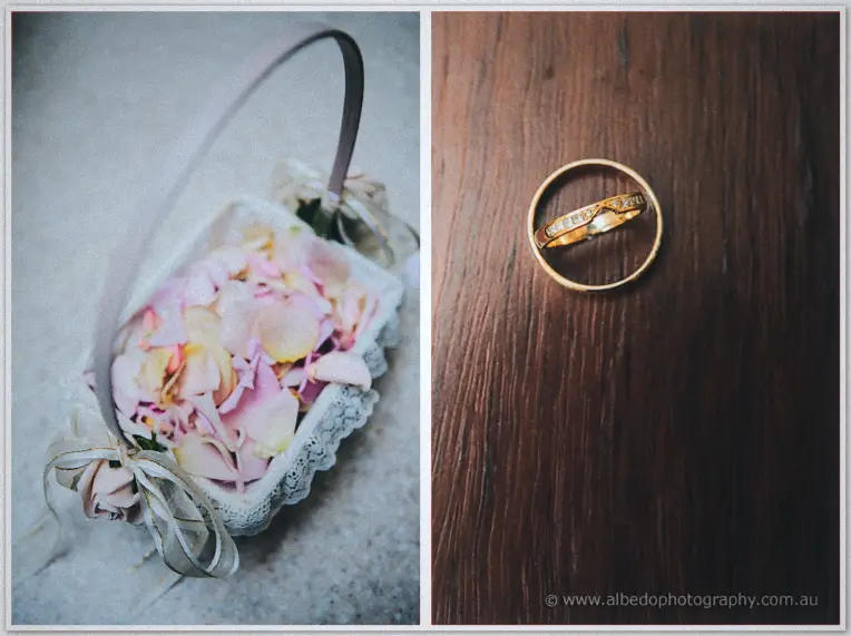 A lace-trimmed basket of pink rose petals sits beside two gold wedding rings on dark timber.