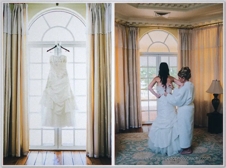 Sarah's gown glows in an arched Brookleigh window before a woman in white helps zip her in.