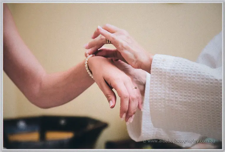 A woman in a white robe clasps a pearl bracelet onto Sarah's outstretched wrist.