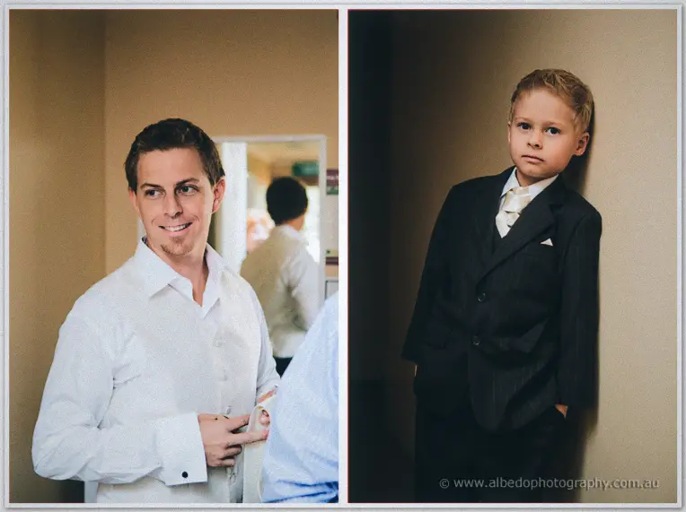 A smiling groom buttons his cuffs beside a page boy in a pinstripe suit leaning on a wall.