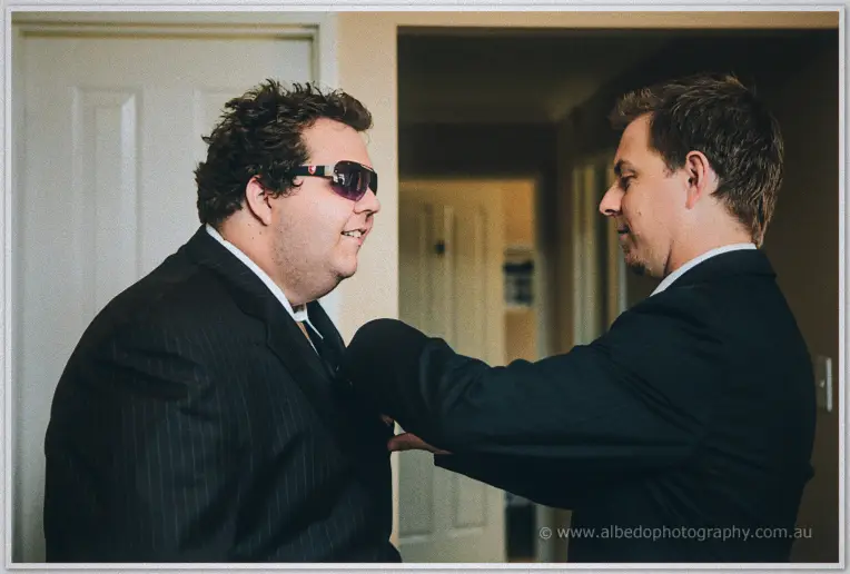 A groomsman in sunglasses laughs as Daniel straightens his pocket square indoors.