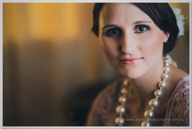 Bridesmaid in mauve lace with a chunky pearl necklace gazes at the camera, white flower in her dark updo.