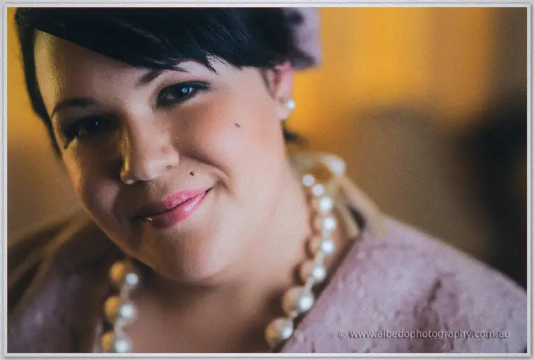 Bridesmaid with cropped dark hair and pearl necklace smiles gently, golden room light glowing behind her.