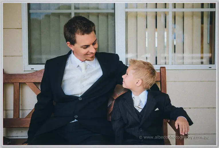 Groom in a white vest shares a smile with his young page boy on a timber bench along the veranda.
