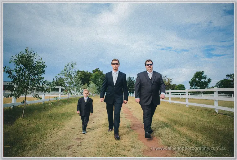 Groom and groomsman walk the dirt path with their pageboy between white paddock fences under a big sky.