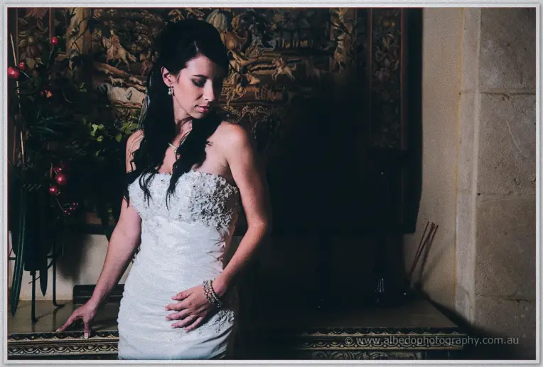 Bride glances down in soft window light, one hand on a carved table beneath a historic tapestry.