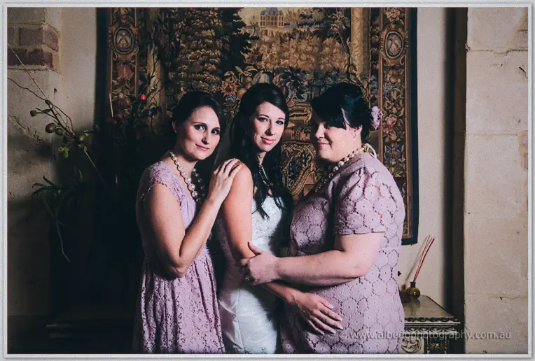 Two bridesmaids in mauve lace flank the bride against a tapestry wall, hands on her shoulder and waist.