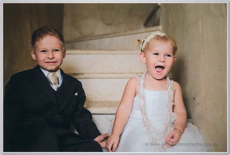 Flower girl laughs mid-giggle beside the pageboy on the stone staircase, hand in hand.