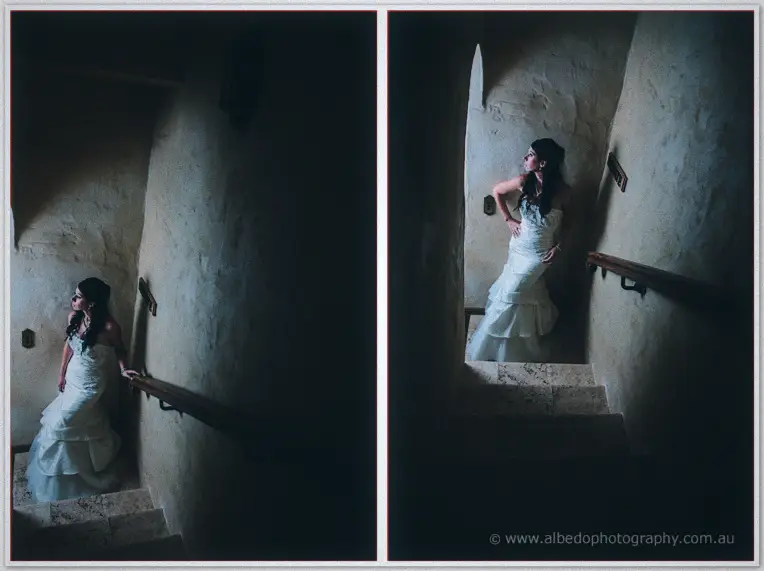 Bride stands on a dim stone stairwell landing, hand on the rail, light falling from a small window.