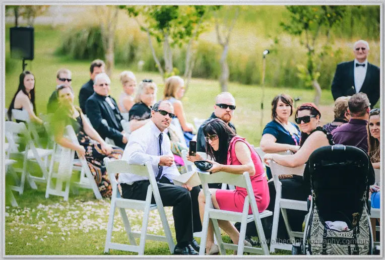 Wedding guests seated on white chairs across the lawn smile toward the ceremony under soft afternoon light.