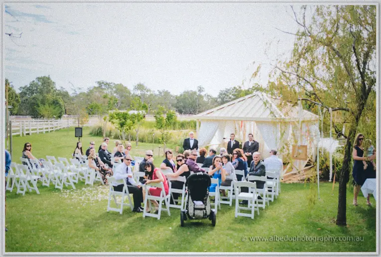 Wide view of the garden ceremony, guests on white chairs facing the draped pavilion by the willow.