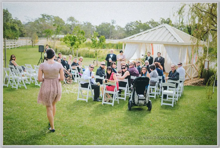 Bridesmaid walks barefoot across the lawn toward seated guests as the groom waits at the pavilion.