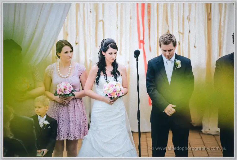 Groom bows his head at the microphone as the bride looks down, bridesmaid and pageboy behind the canopy.