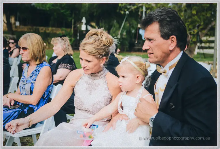 Guests in formal dress smile from the front row, the little flower girl holding a toy on her mother's lap.