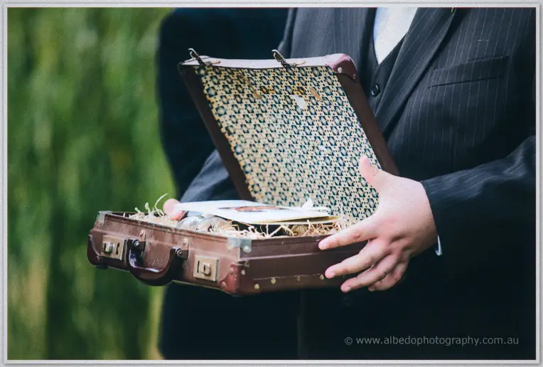 Close-up of an open vintage suitcase holding the rings, straw and cards, cradled in a groomsman's hands.