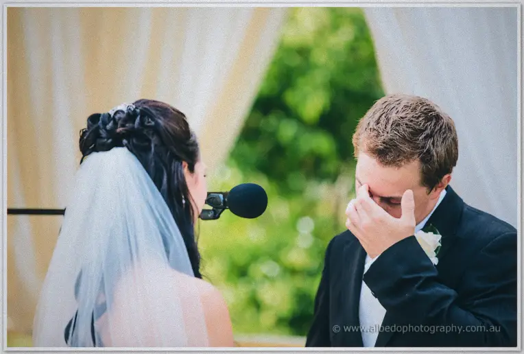 Groom wipes away tears at the microphone while the veiled bride reads her vows in the canopy.