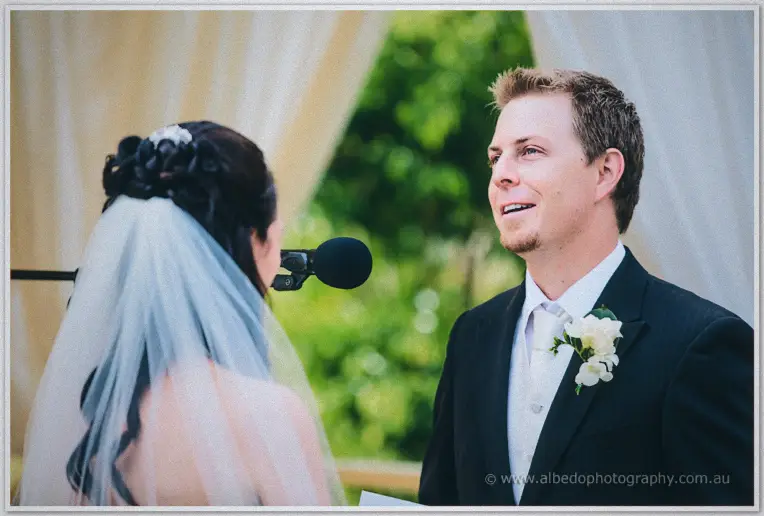 Groom grins up at his bride through the vows, veil and canopy drapes glowing behind them.