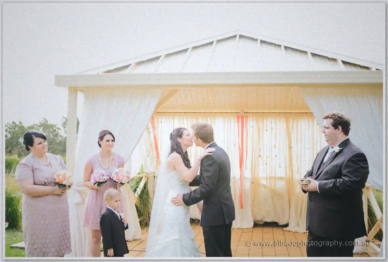 First kiss beneath the white pavilion, bridesmaids and pageboy watching as the celebrant looks on.