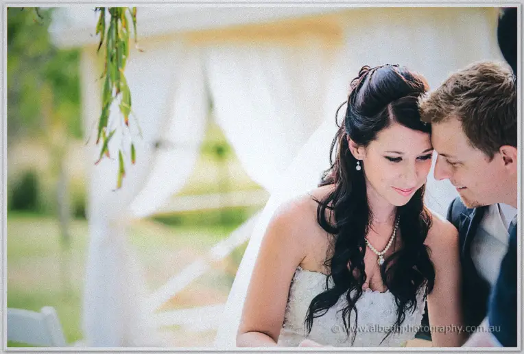 Bride and groom press heads together by the willow, her pearl pendant catching the light.