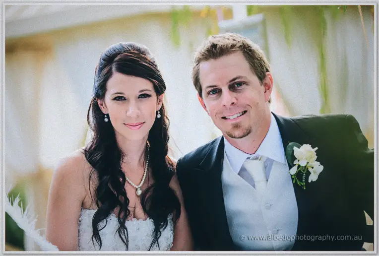 Sarah and Daniel smile side by side at the pavilion, her dark curls beside his white orchid boutonniere.