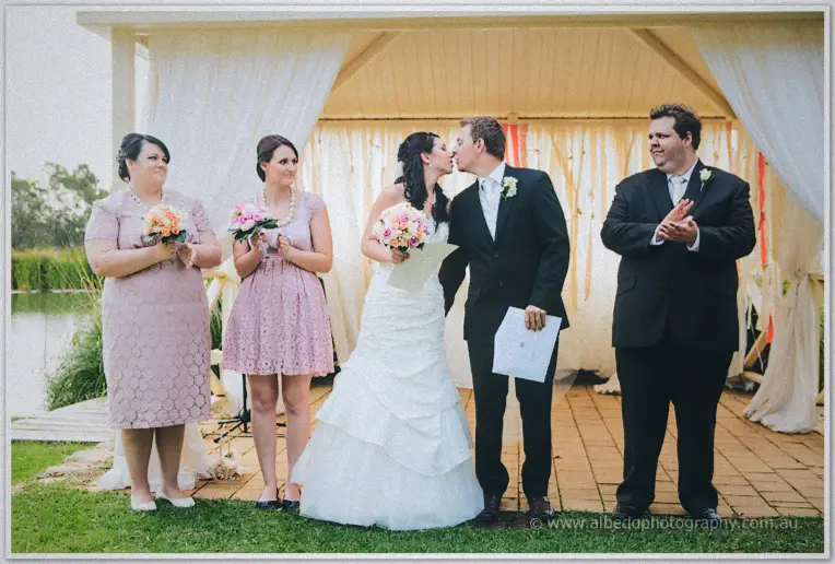Newlyweds kiss on the lawn as bridesmaids laugh and the celebrant applauds beside the lakeside pavilion.