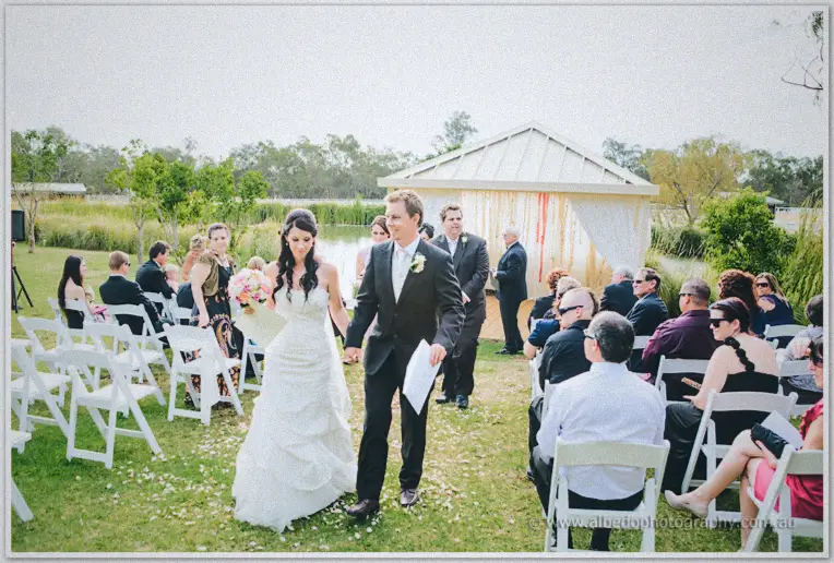 Sarah and Daniel walk the petal-lined aisle hand in hand past cheering guests and the lakeside marquee.