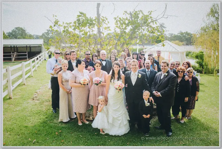 Full wedding party group shot on the lawn between white fences and the homestead stable, soft overcast light.