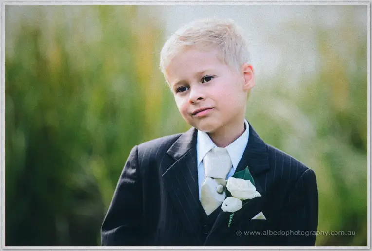 Portrait of the blond pageboy in his pinstripe suit and white rose boutonniere against blurred reeds.