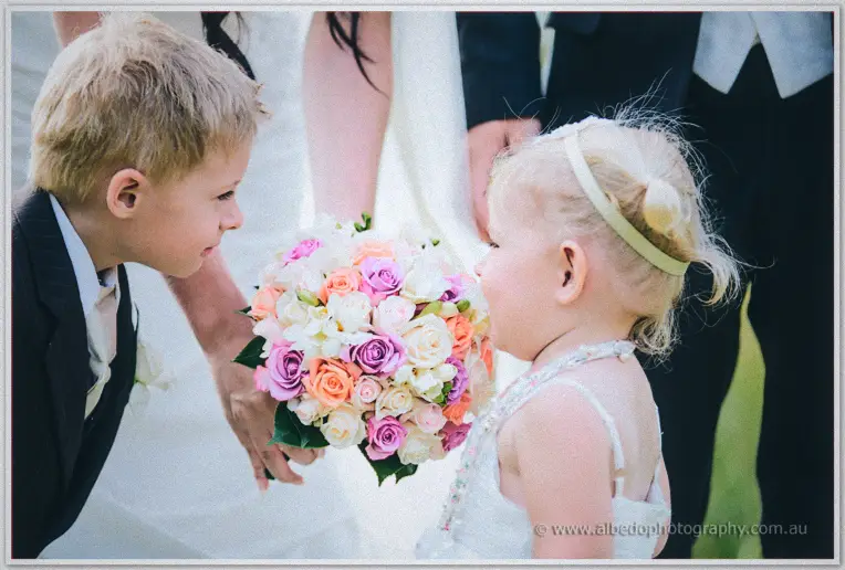 Pageboy and flower girl lean in to smell the bride's pink and peach rose bouquet held low between them.