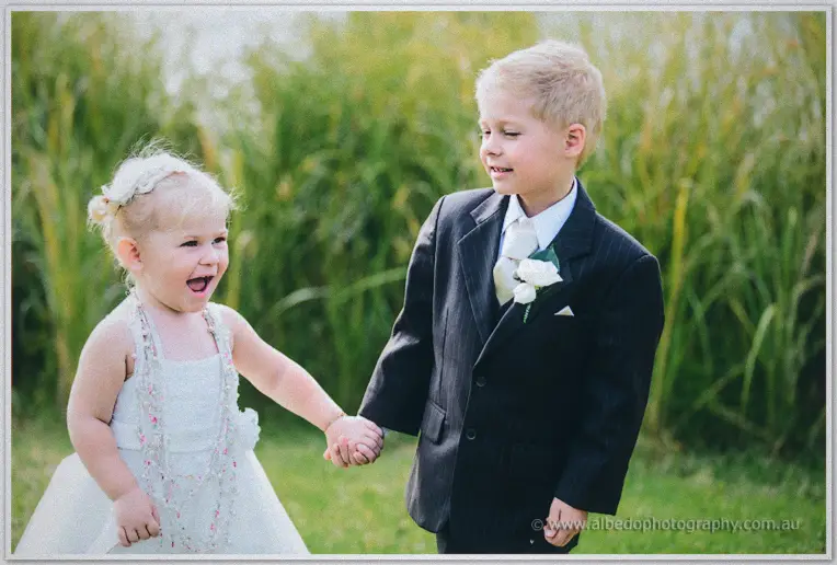 Giggling flower girl clasps the pageboy's hand in the tall grass, his white rose boutonniere bright.