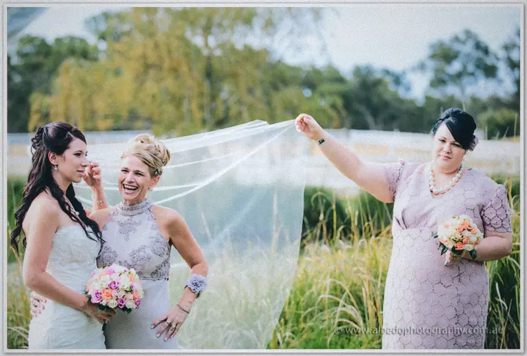 Bridesmaid laughs as she lifts the bride's veil in the breeze beside a friend with peach roses.