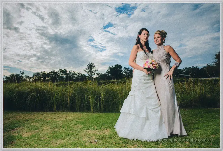 Bride and a guest in champagne lace pose together on the lawn under a dramatic clouded sky.
