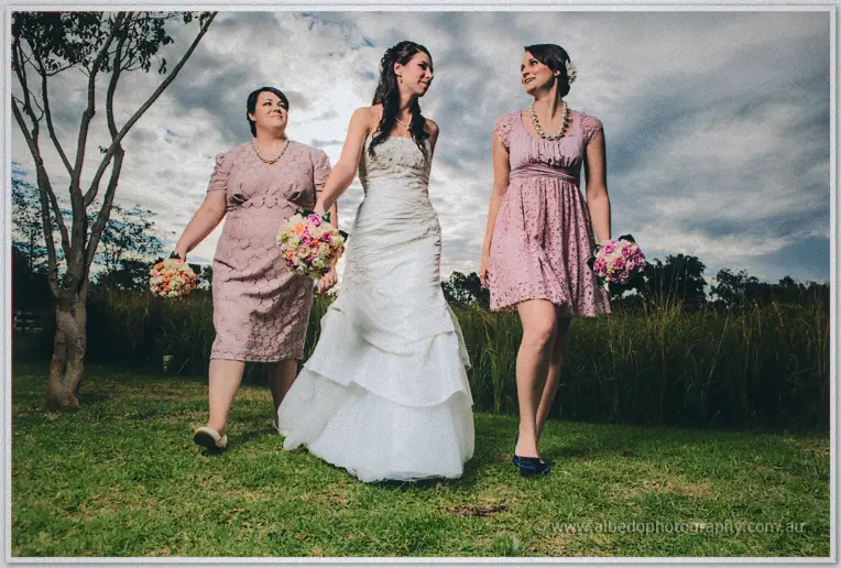 Bride Sarah strolls arm in arm with her two bridesmaids in pink lace through the Brookleigh gardens.