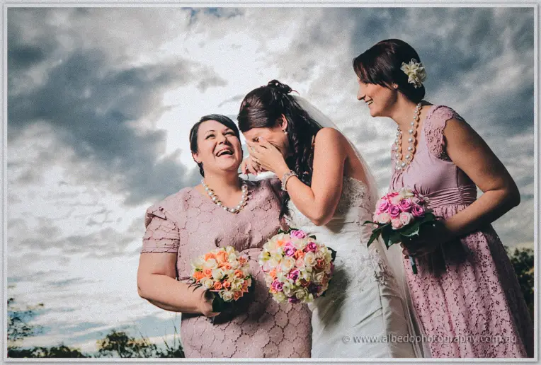 Sarah bursts out laughing as her bridesmaids embrace her beneath a stormy sky at Brookleigh.