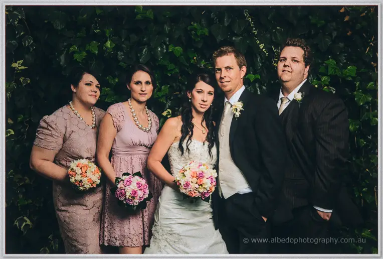 Sarah and Daniel stand with their bridal party before a leafy green wall in dusky light.