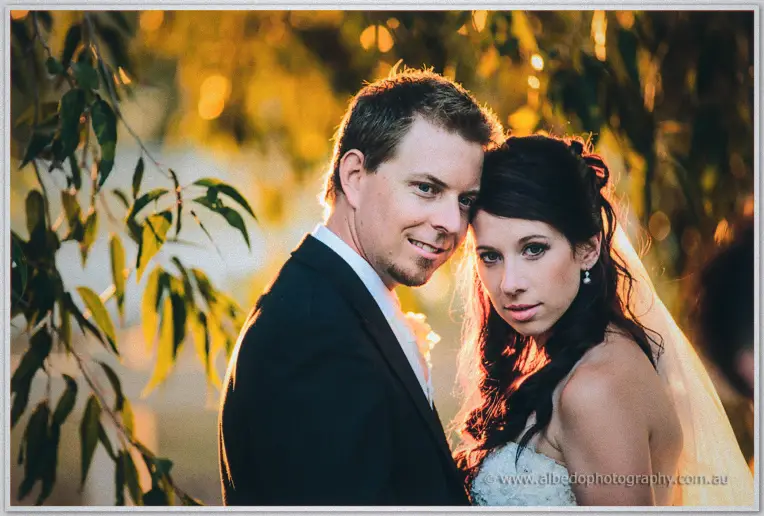 Sarah and Daniel lean close beneath golden leaves as evening light glows around them at Brookleigh.