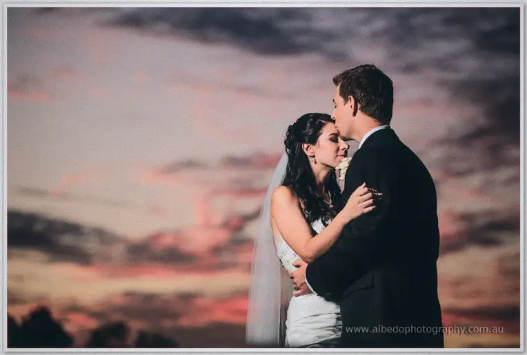 Daniel wraps Sarah in his arms as dusky pink clouds streak the sky above the estate.
