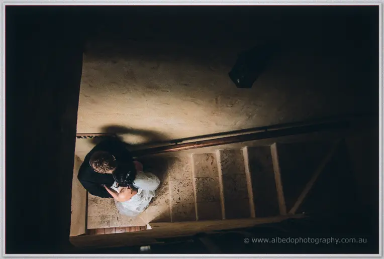 Seen from above, the couple embrace at the foot of a stone stairway in dim lamplight.