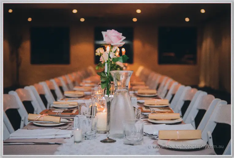 A single pale rose centers the long bridal table, candles flickering between rows of white chairs.