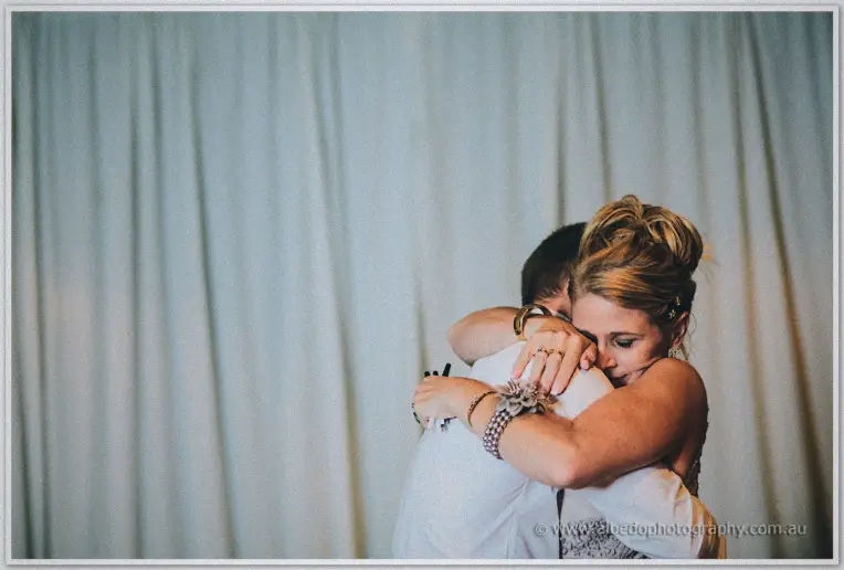A wedding guest in a corsage hugs a friend tightly with eyes closed against white drapery.