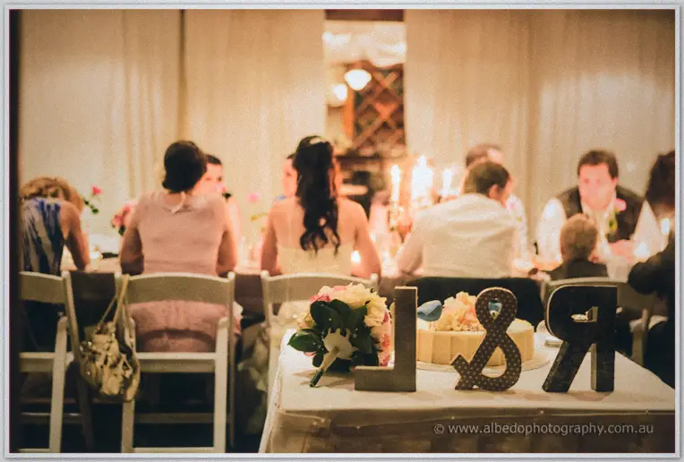 Candlelight washes over seated guests as a monogram, cake and bouquet dress the foreground table.