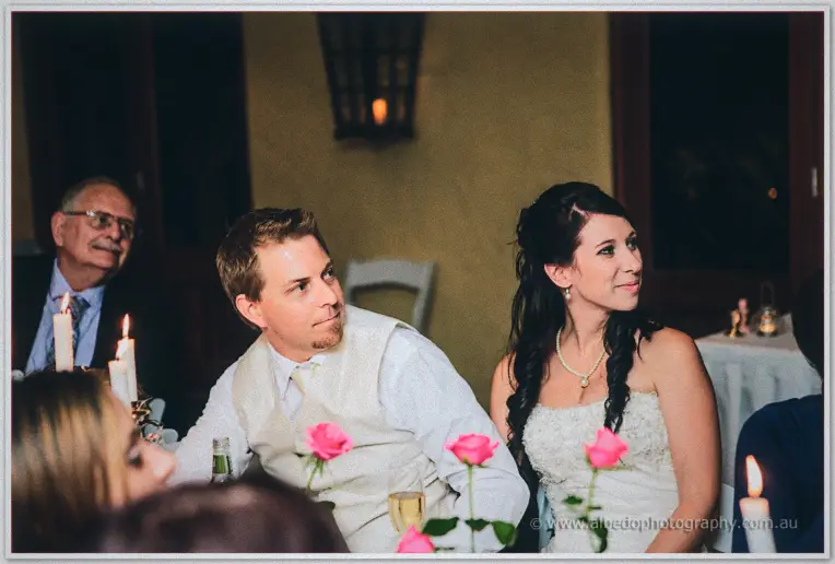 Sarah beams in candlelight as Daniel listens intently to toasts amid pink roses at the head table.
